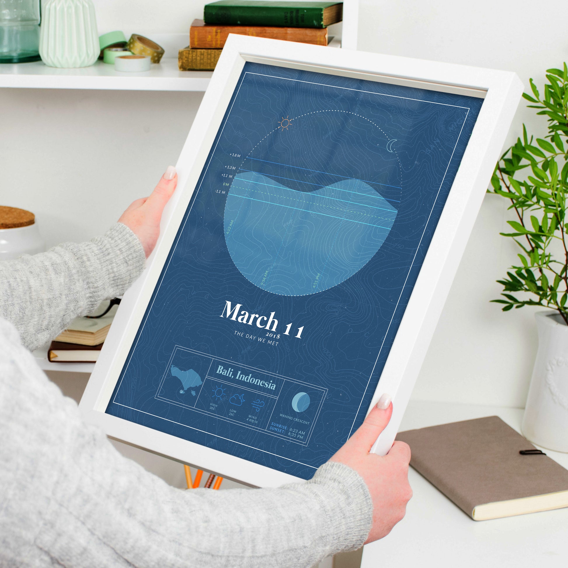 woman holding a white framed picture of the personalized tide poster by salt atlas in the vintage cobalt color in a home setting. These are custom posters showing the tide, weather, and moon phase for a special day, like an anniversary or birthday.