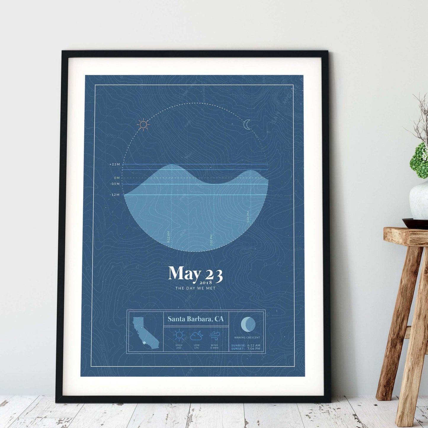black framed picture of the personalized tide poster by salt atlas in the vintage cobalt color in a home setting. These are custom posters showing the tide, weather, and moon phase for a special day, like an anniversary or birthday.